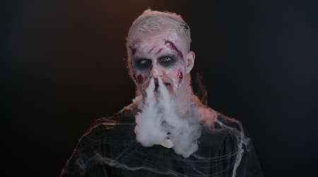 Creepy man with bloody scars face, Halloween stylish zombie make-up. Scary wounded undead guy blows smoke from nose and mouth, smiles terribly. Voodoo rituals. Thematic party. Sinister beast, monsterの写真素材