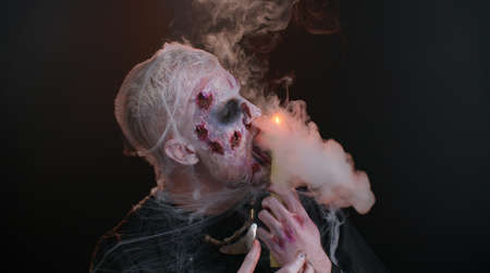Frightening man with Halloween zombie bloody wounded makeup spells conjures over a candle. Horror theme. Sinister undead guy isolated on studio black background. Voodoo rituals. Fashion body artの写真素材