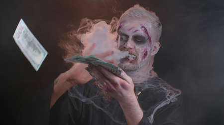 Portrait of sinister man in carnival costume of Halloween crazy zombie with bloody wounded scars face throwing money dollar cash banknotes. Horror theme of cosplay wounded undead, beast, monsterの写真素材