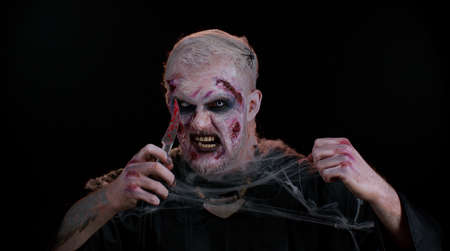 Zombie man with make-up with fake wounds scars and white contact lenses holding bloody knife, trying...