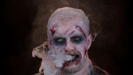 Sinister man with horrible scary Halloween zombie makeup in costume looking ominous at camera blows smoke from nose and mouth. Dead guy with wounded bloody scars face isolated against black backgroundの写真素材