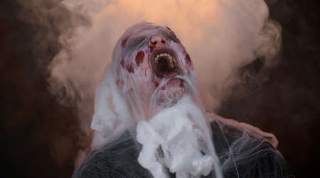 Sinister man with horrible scary Halloween zombie makeup in costume blows smoke from nose and mouth, screaming, shouting, trying to scare, breaks spider web. Dead guy with wounded bloody scars faceの写真素材