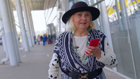 Senior tourist grandmother walking on international airport hall carrying luggage suitcase texting...