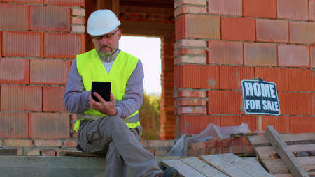 Male construction engineer. Architect with a digital tablet computer at construction site. Man worker foreman looking analyzing blueprints, building house place on background. Construction conceptの写真素材
