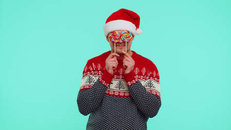 Funny man in red New Year sweater, hat holding candy striped lollipops, hiding behind them, fooling around, dancing isolated on blue studio wall background. Happy Christmas celebration merry holidayの写真素材