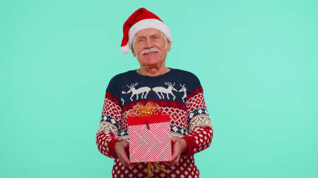 Elderly grandfather man in Christmas sweater, Santa hat getting present gift box, expressing amazement, extreme happiness isolated on blue background. Happy New Year celebration merry holiday conceptの写真素材
