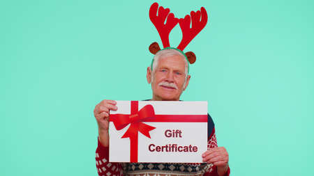 Elderly old grandfather man wears red New Year sweater deer antlers presenting card gift certificate coupon voucher isolated on blue studio wall background. Happy Christmas celebration merry holidayの写真素材
