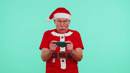 Worried senior grandfather man wears New Year t-shirt enthusiastically playing racing video games on mobile phone isolated on blue studio wall background. Happy Christmas celebration merry holidayの写真素材