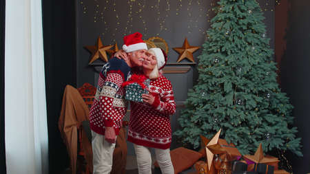 Happy elderly couple in Santa Claus hats celebrating New Year in cozy living room at home, grandfather gifting Christmas present box to surprised grandmother. Mature people celebrate Christmas at homeの写真素材