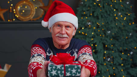 Senior grandfather old man wears red Christmas sweater presenting Christmas gift box stretches out hands in cozy living room at home. Happy mature elderly family New Year Xmas celebration holiday eveの写真素材