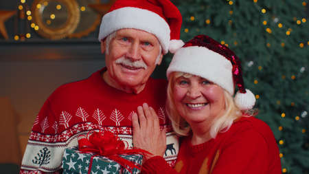 Happy senior couple in Santa Claus hats and sweaters celebrating Christmas in cozy living room at...