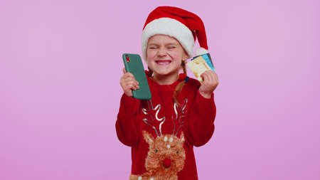 Child girl in Christmas sweater use mobile cell phone, plastic credit bank cards, browsing say wow yes, found out great big win, doing winner gesture. Xmas New Year holidays purchases online shoppingの写真素材
