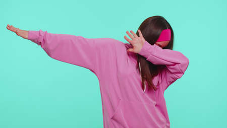 Trendy cheerful positive teen girl having fun dancing and moving to rhythm, dabbing raising hands, making dubdance gesture. Young pretty stylish adult woman. Indoor studio shot on blue wall backgroundの写真素材