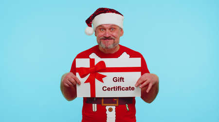 Funny 50s years old man wears red New Year t-shirt and hat presenting card gift certificate coupon voucher isolated on blue studio wall background. Happy Christmas celebration merry holiday conceptの写真素材
