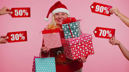 Senior grandmother in Christmas sweater holding many gift boxes and receive discounts inscriptions banner texts. Black Friday. Good holiday low prices for shopping. Blue studio background. New Yearの写真素材