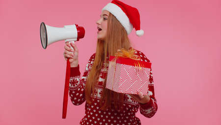 Cheerful woman 20s in red Christmas Santa sweater with gift box scream in megaphone announces discounts sale shopping advertisement on pink background studio. Happy New Year celebration merry holidayの写真素材