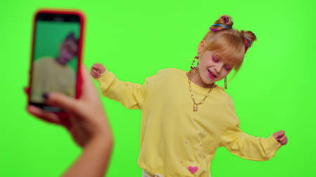 Dancing young little kid girl blogger record dance moves at camera for social media content using phone indoors on chroma key green screen background. Children schoolgirl perform trendy funny danceの写真素材