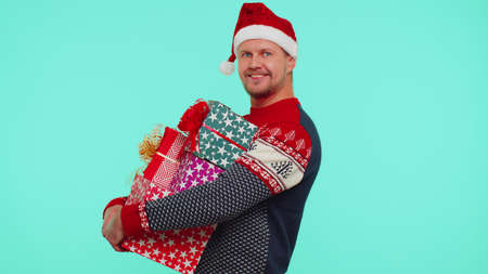 Handsome man in Christmas sweater and hat looking surprised at mount of boxes hands, excited by so many holiday gifts, lot of presents on blue studio background. Happy New Year shopping sale holidaysの写真素材