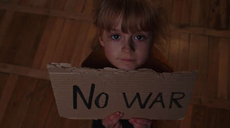 Portrait of afraid poor toddler girl kid homeless sitting at home floor at night holding banner with inscription massage No War. Child alone hiding from bombing attack. Crisis, peace, stop aggressionの写真素材