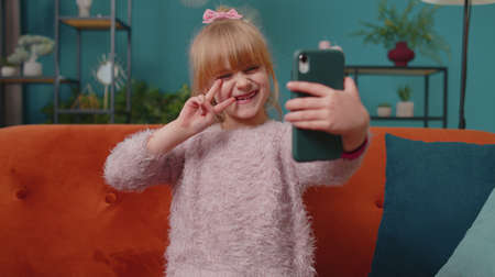 Little elementary toddler school kid girl taking selfie photo with smartphone while sitting on sofa at home. Funny child make blogger self portrait, holding mobile phone in modern flat living roomの写真素材