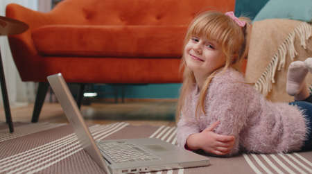 Little school kid girl use laptop computer lying on floor alone at home. Child learning reading...