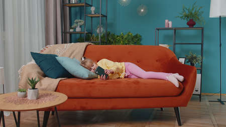 Tired little school kid girl using mobile phone technology device lying on sofa alone at home. Child toddler hold smartphone watching online cartoons, chatting with friends. Children tech addictionの写真素材