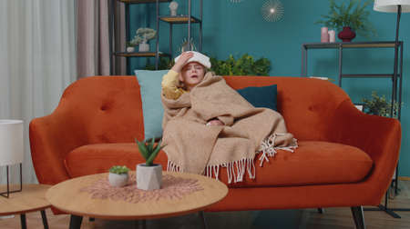 Portrait of one Caucasian child girl suffering from cold or allergy in plaid sitting on sofa in living room at home. Sick school kid with towel on head blows nose snot into napkin. Coronavirus conceptの写真素材