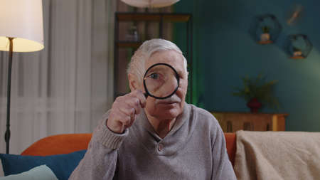 Senior one grandfather man looking through magnifying glass as if reading, researching, investigating, examining, analyzing something. Mature old granpa alone sitting on sofa at home looking at cameraの写真素材