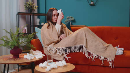 Young woman feels sick measuring body temperature with electronic thermometer at home alone. High fever. Concept of healthcare and illness. Coronavirus quarantine, pandemic or seasonal grippe symptomsの写真素材