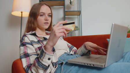 Smiling Caucasian young woman lying on sofa using credit bank card and laptop computer while transferring money, purchases online shopping in living room home. Female girl tapping, scrolling, browsingの写真素材