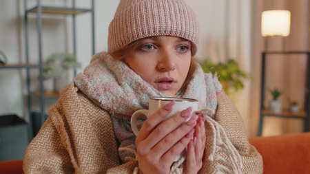 Young sick woman wear hat wrapped in plaid sit alone shivering from cold on sofa drinking hot tea in...