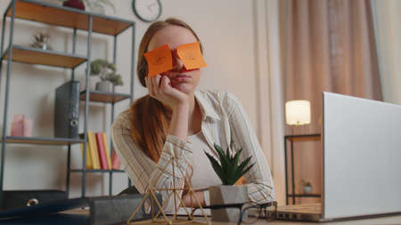 Tired woman freelancer using laptop computer napping at home office desk with funny stickers on eyes...
