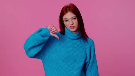 Upset pretty stylish teen girl in blue sweater showing thumbs down sign gesture, expressing discontent, disapproval, dissatisfied, dislike. Young adult woman. Indoor studio shot on pink backgroundの写真素材