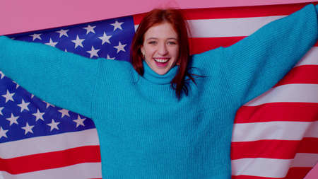 Trendy cheerful positive teen girl waving and wrapping in American USA flag, celebrating, human rights and freedoms. Independence day. Young pretty adult woman. Indoor studio shot on pink backgroundの写真素材