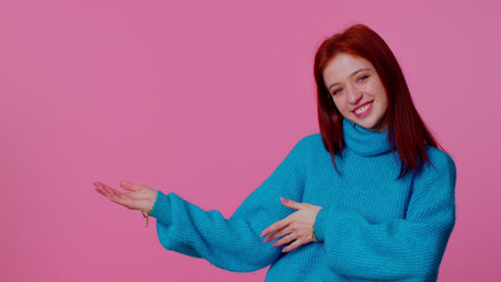 Pretty red hair teenager girl showing thumbs up and pointing empty place, advertising area for commercial text, copy space for goods promotion. Young adult woman. Studio shot on pink wall backgroundの写真素材
