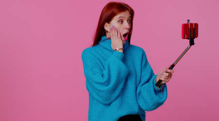 Wow reaction. Impressed shocked teenager student adult girl blogger taking selfie on mobile phone selfie stick, communicating video call online. Young fashionable woman on pink studio backgroundの写真素材