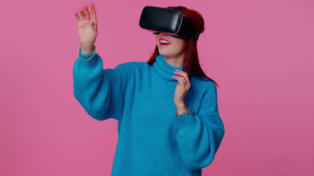 Teen redhead girl using virtual reality futuristic technology VR app headset helmet to play...