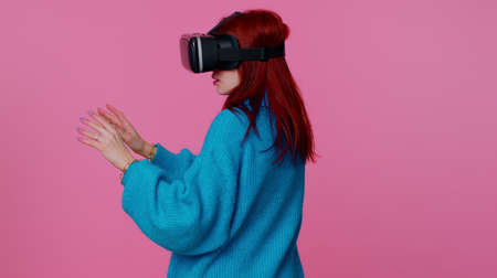 Amazed teen student girl using headset helmet app to play simulation game. Watching virtual reality 3D 360 video. Young woman in VR goggles isolated on pink background. Addiction from technologyの写真素材