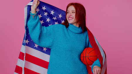Redhead happy young woman 20s sportswoman basketball fan in blue sweater holding American USA flag doing winner gesture, dancing isolated on pink background. Workout sport motivation lifestyle conceptの写真素材