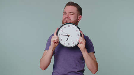 Joyful young man holding clock watch, hiding, checking time on clock, running late to work, being in delay, deadline on pink studio background. Serious guy looking at camera worrying to be punctualの写真素材