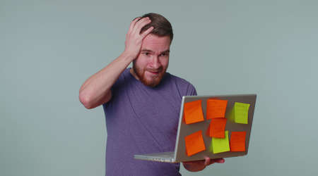 Exhausted man freelancer with pasted sticker notes using laptop computer having concentration problem creative crisis. Nervous adult guy confused by big routine work loading. Gray studio backgroundの写真素材