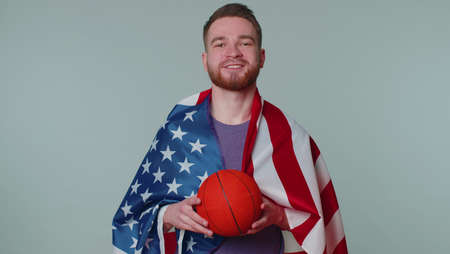 Bearded happy young man 20s sportsman basketball fan in purple t-shirt holding American USA flag doing winner gesture, dancing isolated on gray background. Workout sport motivation lifestyle conceptの写真素材