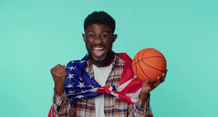 African american one young man 20s sportsman basketball fan in checkered shirt holding American USA flag doing winner gesture, dancing isolated on blue background. Workout sport motivation lifestyleの写真素材