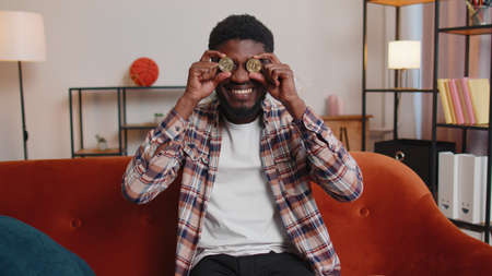 Man freelancer holding two gold coins in living room, successful developer programmer. Guy stock trader earning bitcoins after online monitoring trading operation. Increasing wealth, mining at homeの写真素材
