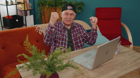 Senior old grandfather man in stylish cap playing computer video games on laptop computer, wins,...