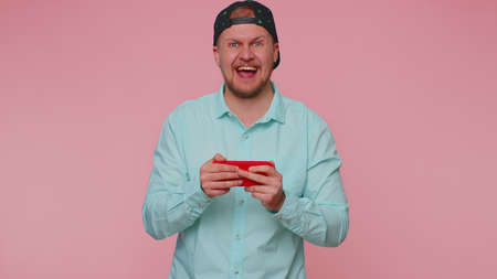 Worried funny addicted man in shirt enthusiastically playing racing video games on mobile phone. Young adult guy using smartphone gadget app with drive simulator isolated on pink studio backgroundの写真素材