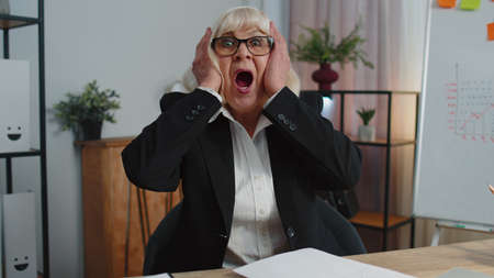 Oh my God Wow. Senior mature older business woman raising hands in surprise looking at camera with big eyes, shocked by sudden victory, good win news at home office. Elderly grandmother freelancerの写真素材