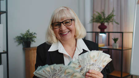 Senior mature banker business woman accountant wears glasses, formal suit, celebrating business success dancing with stack of money dollar cash. Elderly grandmother freelancer indoors office interiorの写真素材