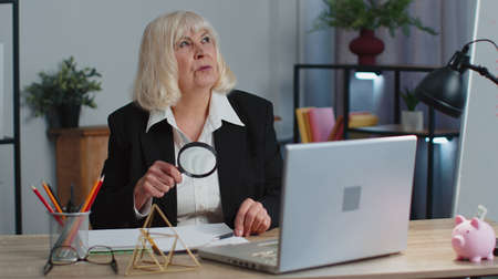 Accountant senior business woman looks through magnifying glass, investigate, examine analyzing reading documents, sits at workplace desk. Elderly grandmother freelancer working at office. Paperworkの写真素材