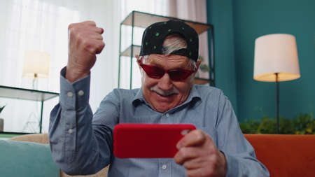 Worried funny senior old grandfather man enthusiastically playing shooter online video games on...